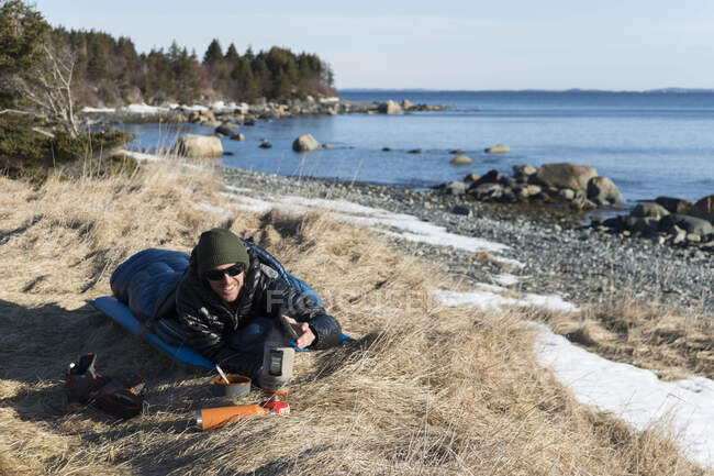 Escursionista maschio preparare il cibo mentre sdraiato nel sacco a pelo sul campo dal mare — Foto stock