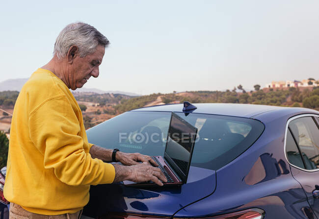 Vista laterale di un anziano viaggiatore che naviga con un computer portatile nel bagagliaio di un moderno veicolo blu in campagna. — Foto stock