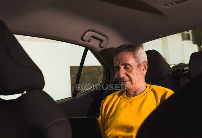 Front view of an elderly traveler sitting in the back seat and typing on a laptop keyboard during a road trip in the countryside — Stock Photo