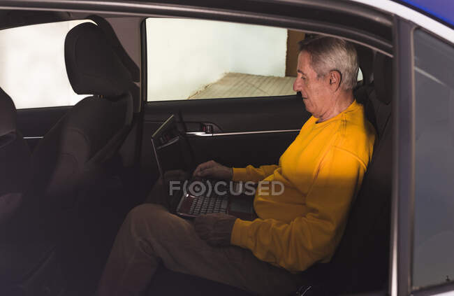 Side view of aged male traveler sitting on back seat and typing on laptop keyboard during road trip in countryside — Stock Photo