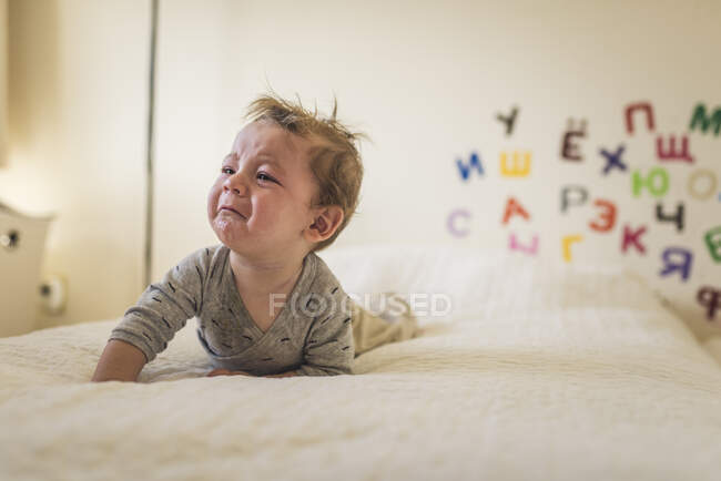 Плачущий ребенок на кровати с белым одеялом и алфавитом на стене позади него — стоковое фото