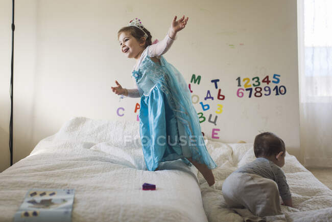 Happy girl wearing princess costume jumping on bed with baby brother — Stock Photo