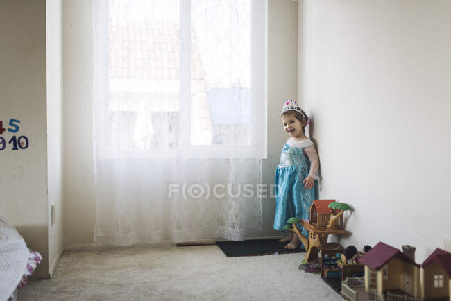 Fille souriante en costume de princesse debout à côté de jouets par la fenêtre — Photo de stock