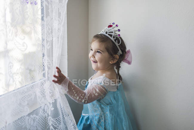 Smiling girl in princess costume and tiara touching white lace curtain — Stock Photo