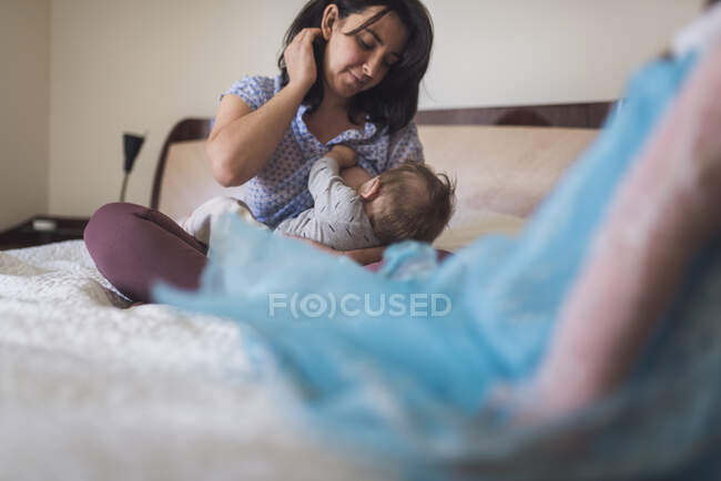 Любить мать 30-ти лет, сидящую на кровати и кормящую грудью ребенка — стоковое фото
