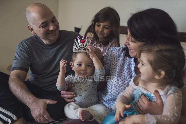 Toda la familia en la cama de los padres riéndose de un bebé de un año con tiara - foto de stock