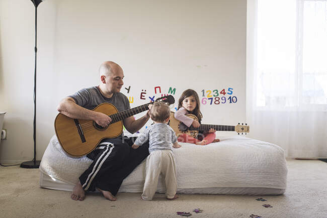 Papá y su hija tocando la guitarra en la habitación de los niños con 1 año de edad viendo - foto de stock