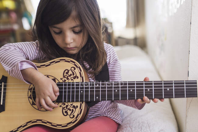 Серйозна 6-річна дівчина, яка вибирає гітарну струну, сидячи на ліжку — стокове фото