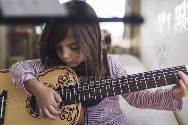 Орієнтована молода дівчина вчиться грати на гітарі, сидячи на ліжку — стокове фото