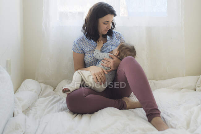 Босоногая мать кормит ребенка грудью на белой кровати перед окном — стоковое фото