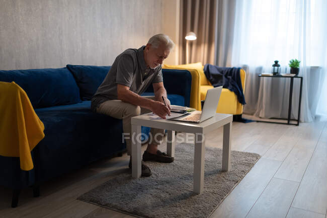 Homem idoso sentado no sofá e checando faturas perto do laptop à noite em casa — Fotografia de Stock