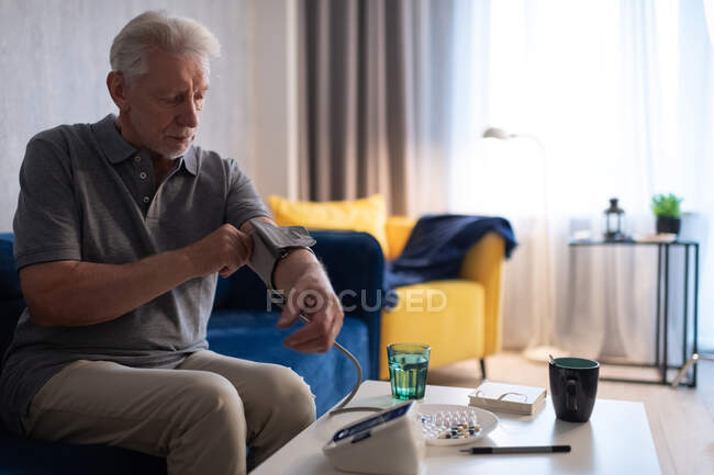 Elderly man putting on cuff of blood pressure monitor on arm on sofa at home — Stock Photo
