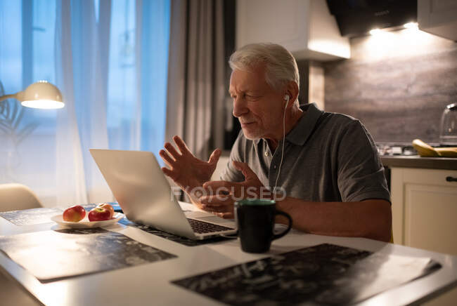 У віці чоловік жестикулює і розмовляє з сім'єю на ноутбуці ввечері на кухні — стокове фото