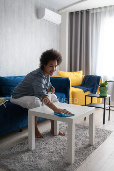 Щаслива етнічна жінка розпилює і протирає стіл, сидячи на дивані вдома — стокове фото