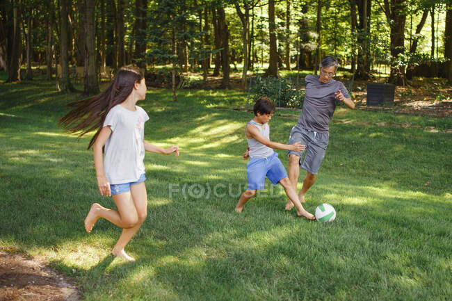 Un papá juega al fútbol descalzo en el patio con sus dos hijos - foto de stock