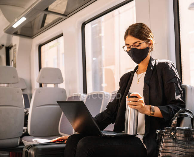 Femmina manager in maschera con thermos e computer portatile di navigazione durante il pendolarismo al lavoro in treno durante la pandemia — Foto stock