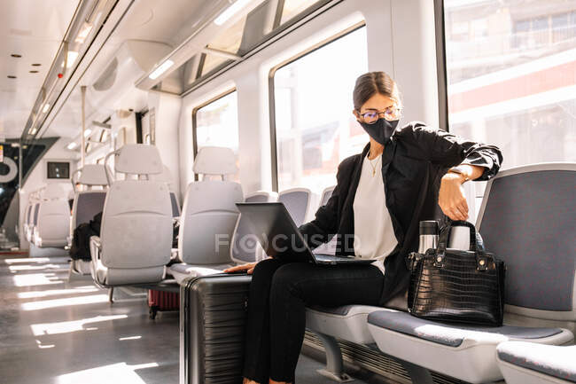 Female manager in face mask holding thermos and browsing laptop while commuting to work by train during pandemic — Stock Photo