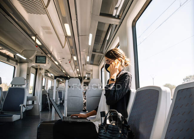 Vista laterale della donna d'affari in maschera di navigazione laptop e rispondere alla telefonata durante la guida del treno moderno al lavoro durante l'epidemia — Foto stock