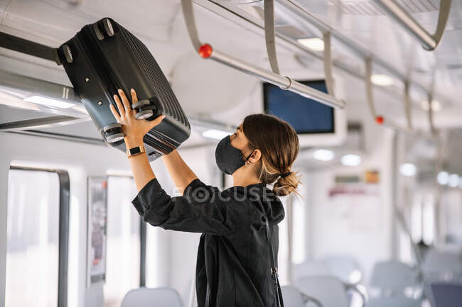 Side view of female manager in fabric mask putting luggage on shelf while commuting to work by train — Stock Photo