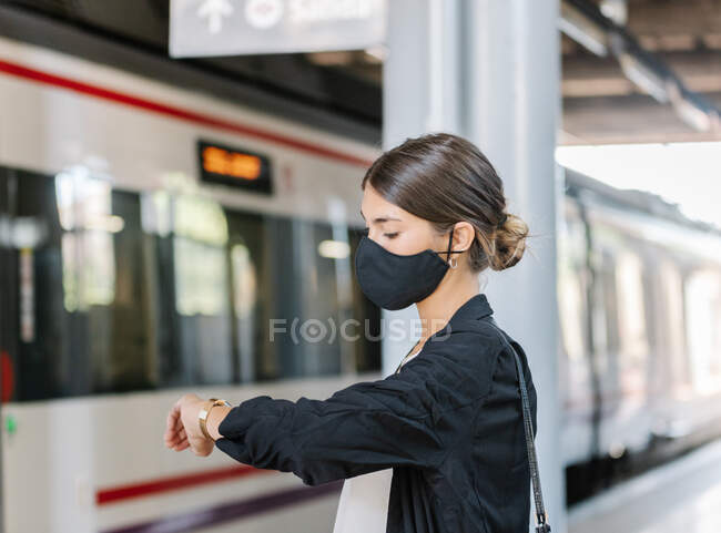 Vista laterale della donna d'affari in maschera che controlla l'orologio da polso mentre si trova vicino al treno sulla stazione ferroviaria durante l'epidemia — Foto stock
