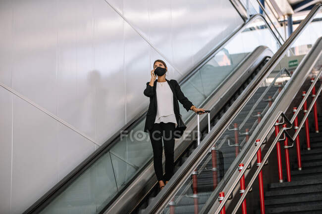 Front view of a woman with suitcase, smartphone and hat riding escalator on station during summer vacation — Stock Photo