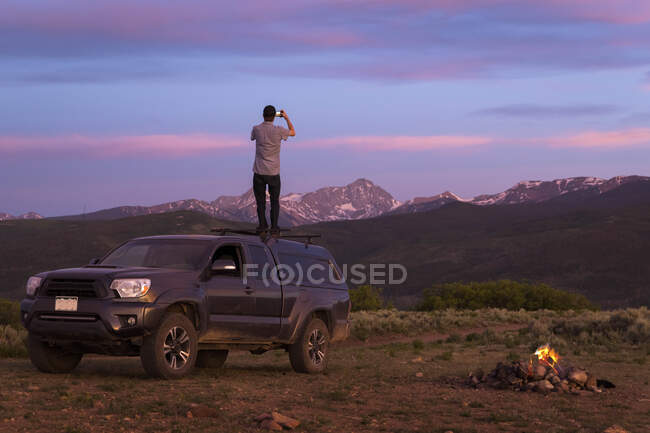 Visão traseira do homem fotografando em pé no carro off-road durante o pôr do sol — Fotografia de Stock