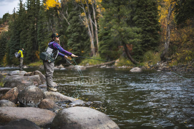 Vista lateral de la pesca con mosca mujer mientras está de pie en la roca en el río Roaring Fork - foto de stock