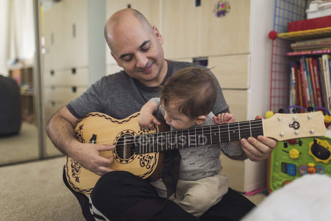 1 yr old playing child's guitar while sitting on bald father's lap — Stock Photo