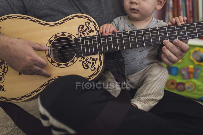 Dad on floor playing child-size guitar while holding 1 yr old on lap. — Stock Photo