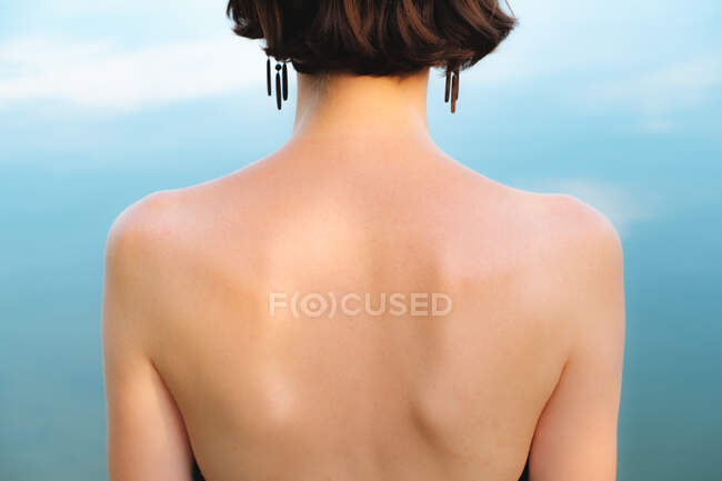 Rücken einer Frau vor blauem Hintergrund. Symmetrie, natürliches Schönheitskonzept: Mittelteil einer unbekleideten jungen Frau vor klarem Wasser, das den Himmel reflektiert — Stockfoto
