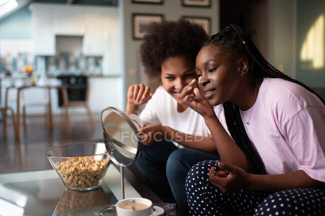 Multiethnic girlfriends with under eye pads smiling and looking at mirror at home — Stock Photo
