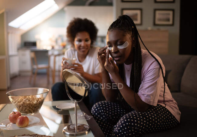 Joven mujer negra aplicando bajo parche ocular durante la pijamada con un amigo - foto de stock