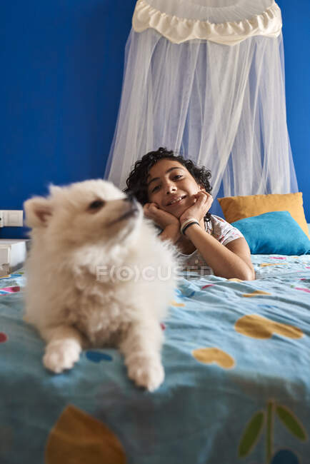 Uma menina e seu cachorro branco em primeiro plano sentado na cama de frente para a câmera. Conceito de cão — Fotografia de Stock