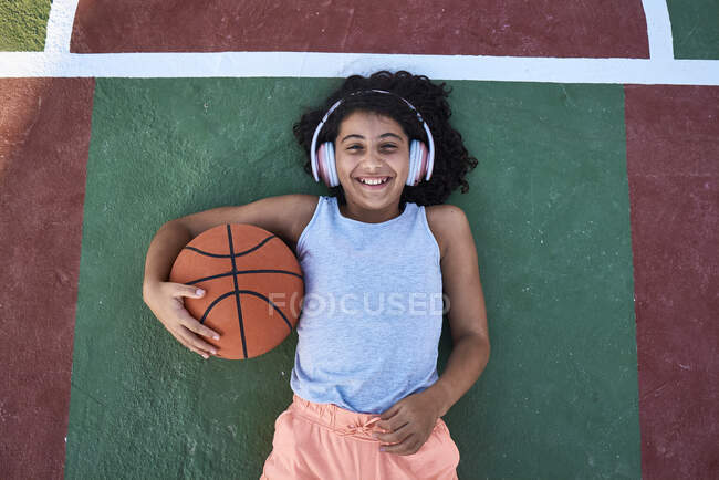 Una niña con el pelo rizado está tirada en una cancha de baloncesto riendo. Concepto de estilo de vida - foto de stock