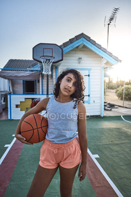 Una chica con el pelo rizado de pie en la cancha de baloncesto de su propia casa. Concepto de estilo de vida - foto de stock