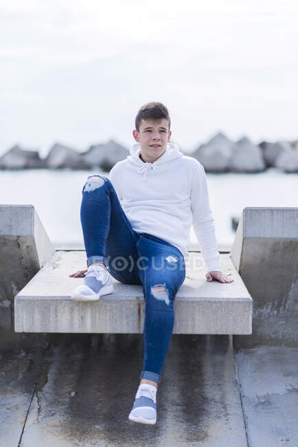 Vista frontal de un adolescente con atuendo casual mientras está sentado en un banco al aire libre y mirando hacia otro lado - foto de stock