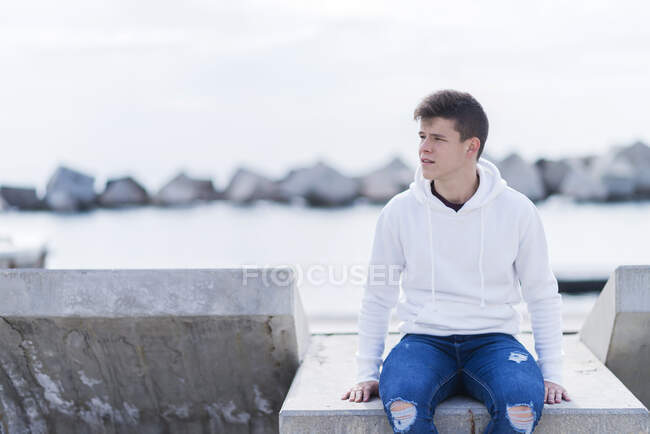 Vista frontal de un adolescente con atuendo casual mientras está sentado en un banco al aire libre y mirando hacia otro lado - foto de stock