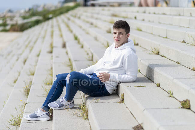 Vista lateral del joven sentado en la escalera al aire libre, mirando hacia otro lado - foto de stock