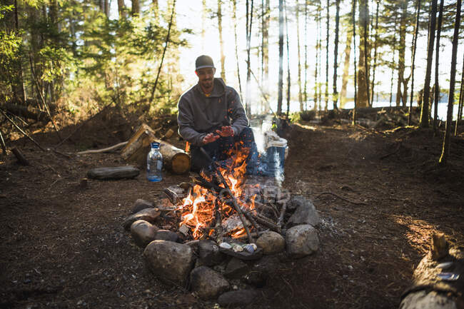 Mann wärmt sich bei Sonnenuntergang am Feuer auf, während er im Küstenstaat Maine campt — Stockfoto