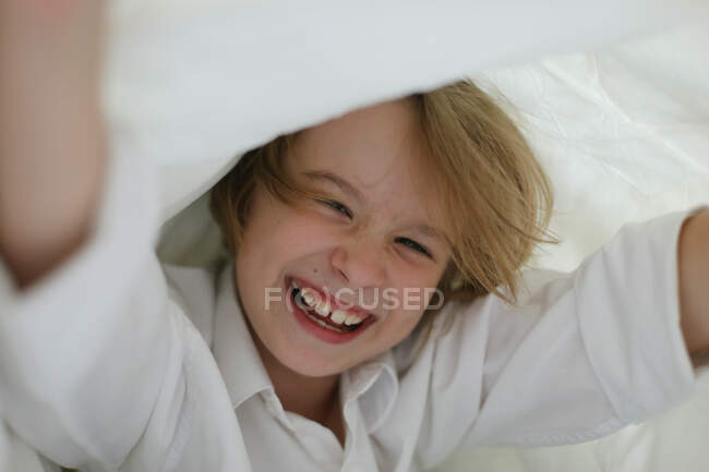 Un bambino con una camicia bianca si diverte sotto le coperte. — Foto stock