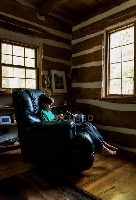 Kleiner Junge liest in Ledersessel in rustikalem Blockhaus. — Stockfoto