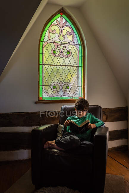 Kleiner Junge liest in Ledersessel vor verziertem Fenster des Hauses. — Stockfoto