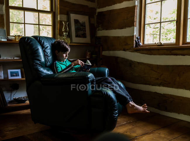 Young boy reading in leather recliner chair in rustic log cabin home. — Stock Photo