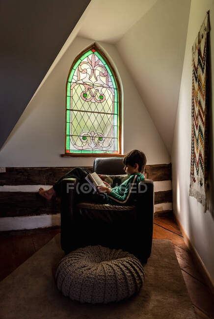 Young boy reading in leather chair in front of ornate window of home. — Stock Photo