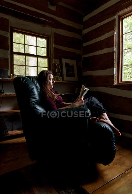 Mulher lendo em cadeira reclinável de couro em uma cabine rústica. — Fotografia de Stock