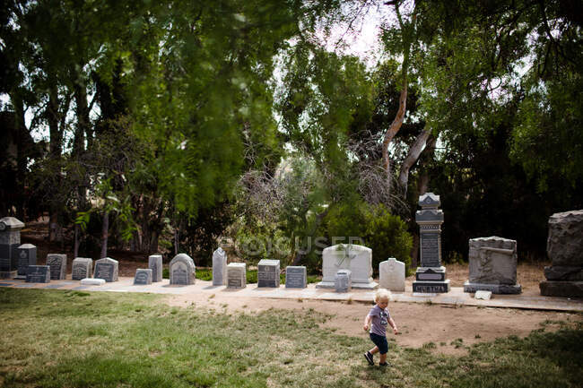 Un enfant de deux ans traverse le parc avec des pierres tombales en arrière-plan — Photo de stock