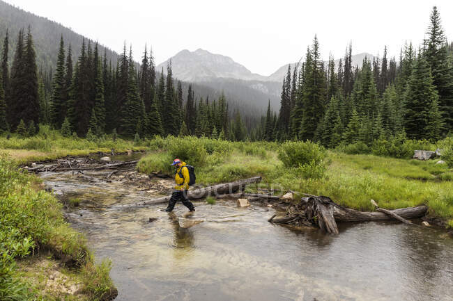 Side view of man hiking in mountains and crossing river on overcast day in British Columbia — Stock Photo