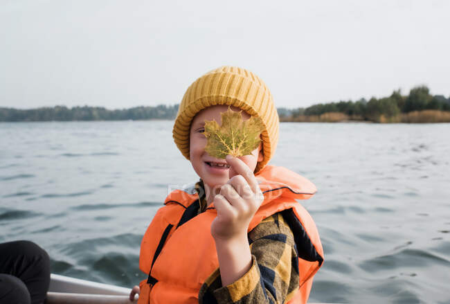 Молодий хлопчик на рибальському човні тримає кленовий лист вгору, виглядаючи щасливим — стокове фото