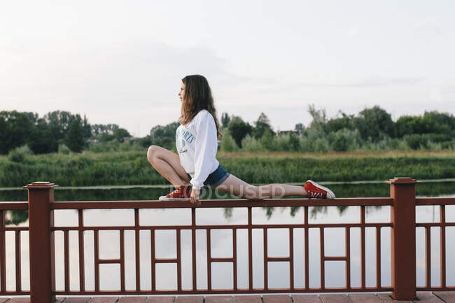Femme étire ses jambes sur une clôture près de la rivière — Photo de stock