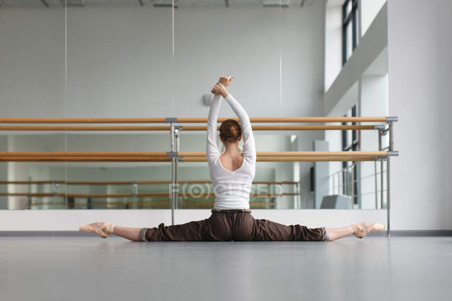 Jeune ballerine en tenue de répétition assise en split près du miroir avec barre, cours de chorégraphie, vue de dos — Photo de stock